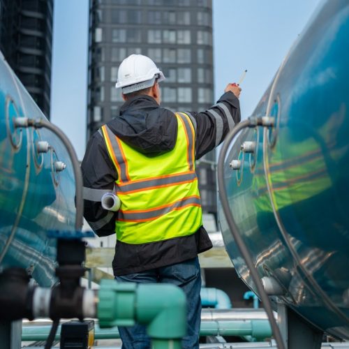 A focused engineer in a reflective vest and hard hat uses a laptop on an urban industrial rooftop, inspecting equipment.